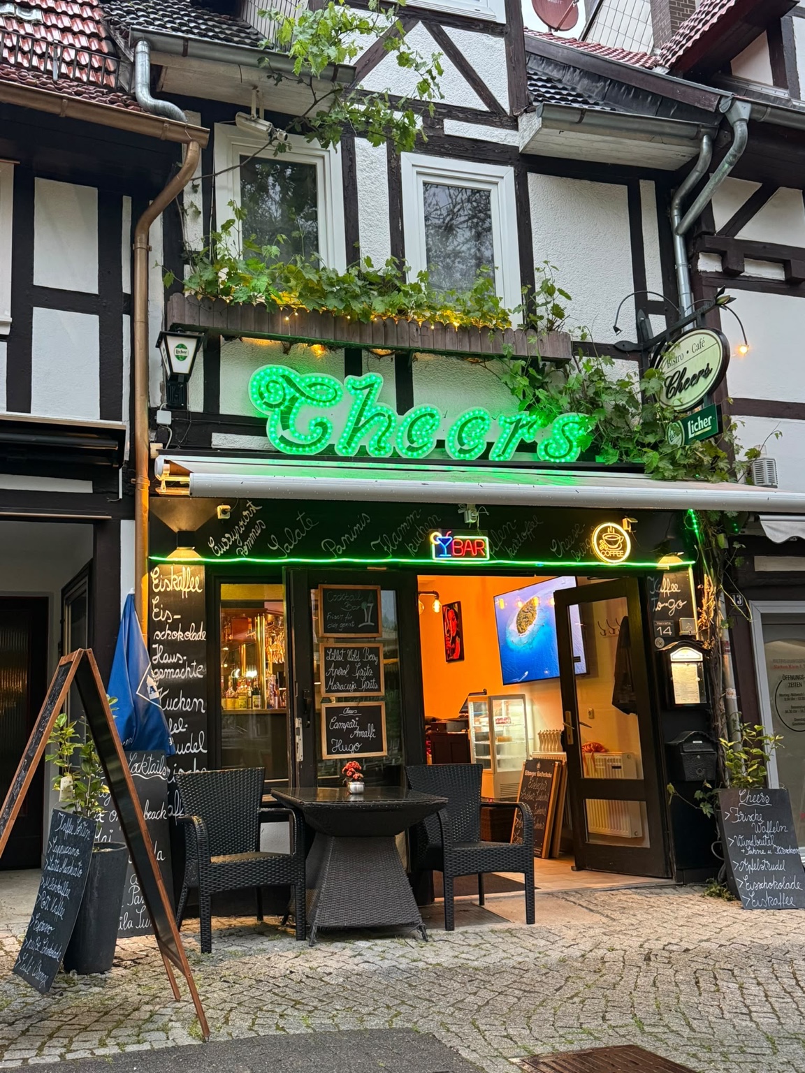 Der Cheers-Eingang im Fachwerkhaus mit grünem Leuchtschriftzug und Sitzplätzen draußen